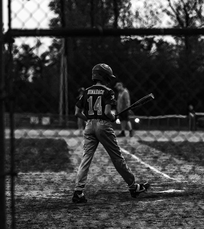 gallery-6 Black and white photo of a young baseball player holding a bat at a game.