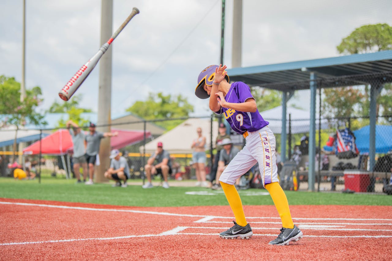 services-02 A young baseball player wearing a helmet dodges a pitch on a sunny day during a game.