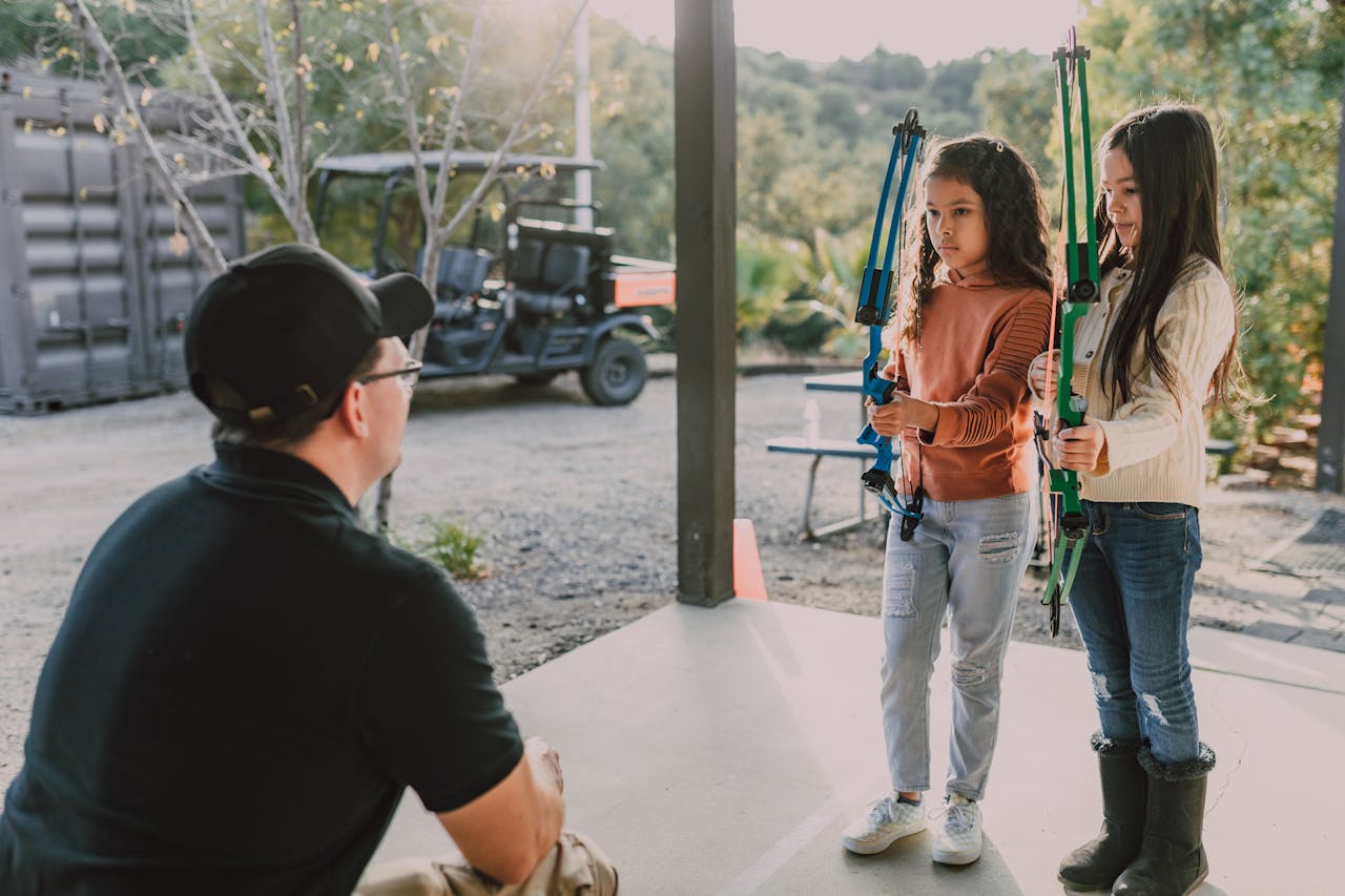 gallery-4 Two young girls learning archery with an instructor outdoors, focused and attentive.